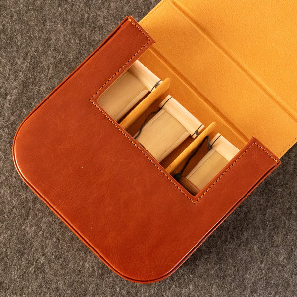 A close-up of an open dudukhouse reed leather case in warm brown and tan, holding three duduk reeds, set on a dark gray textured surface.