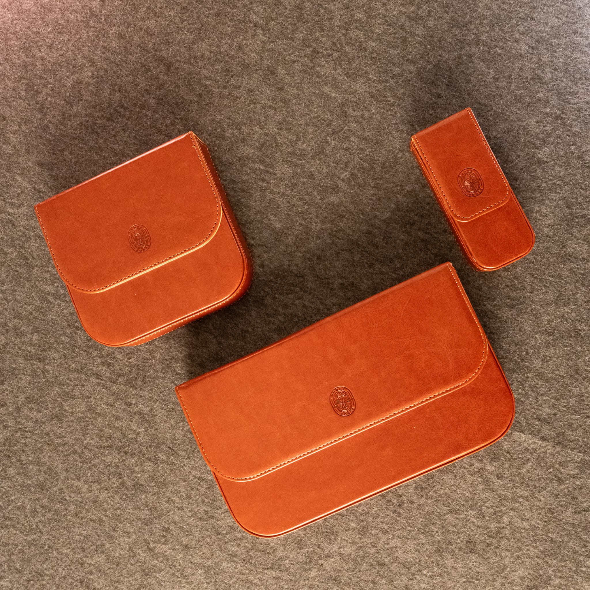 Three orange-brown leather duduk reed  cases made by Dudukhouse in different sizes arranged on a textured gray felt background.