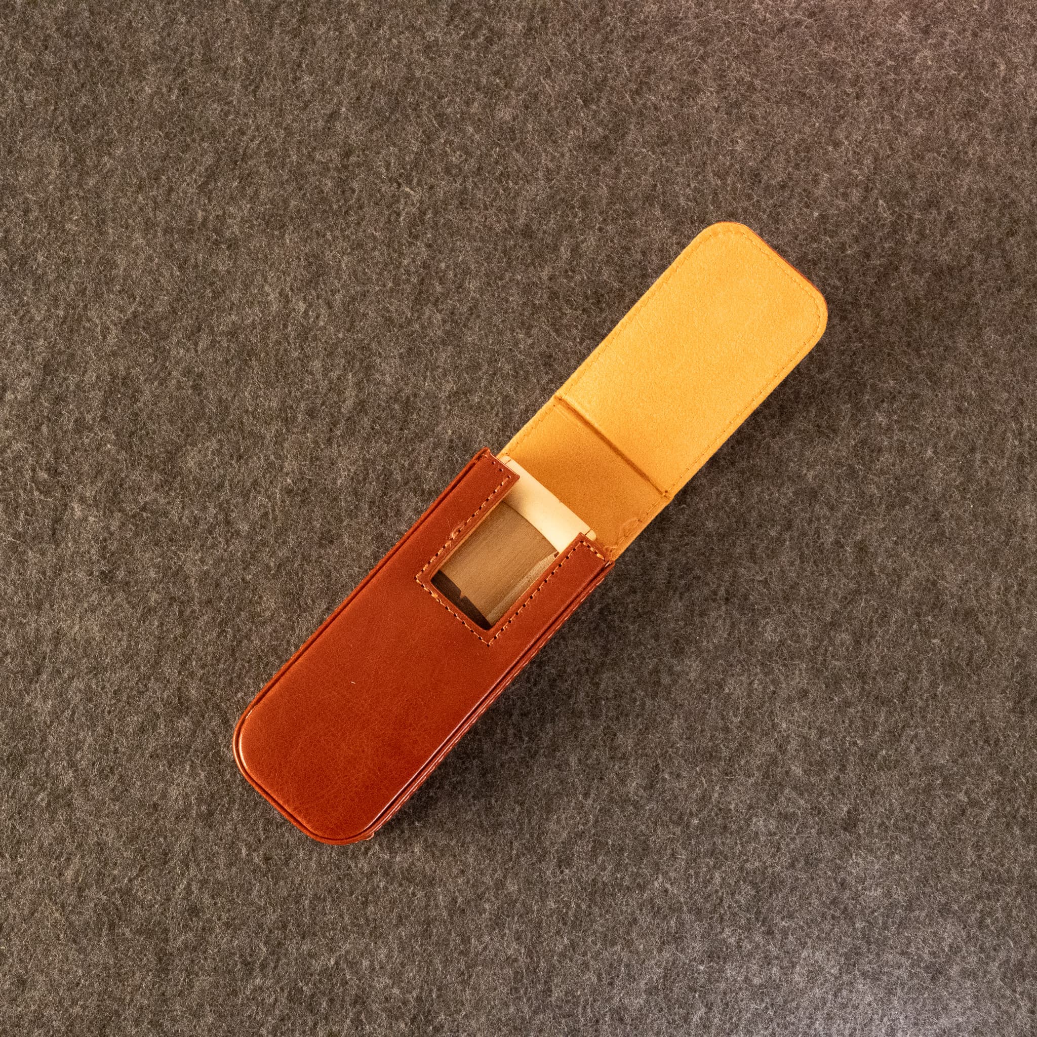 A leather duduk reed case by Dudukhouse, shown open on a textured gray felt background. The exterior is a warm reddish-brown leather, while the interior lining and the visible wooden insert are a light tan.