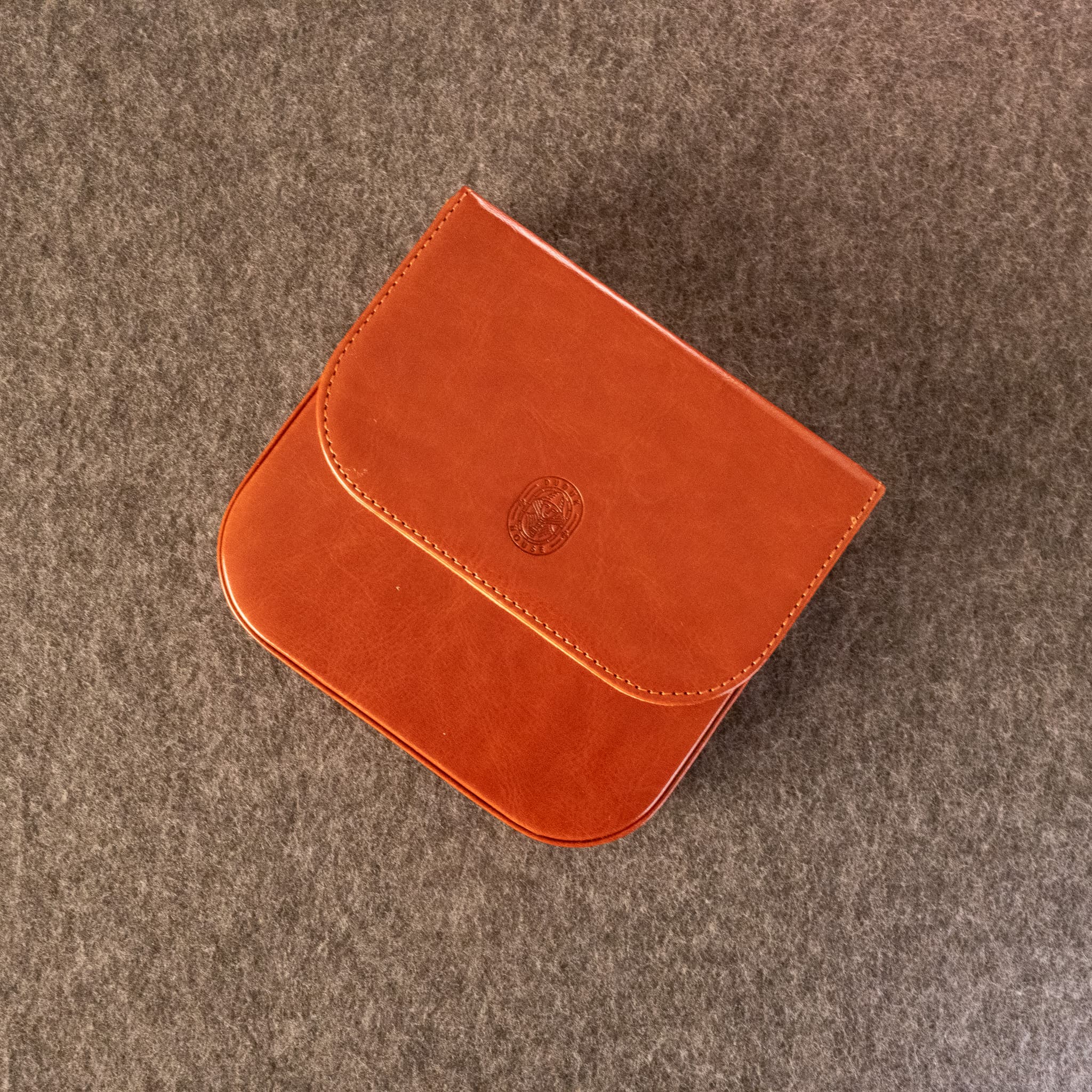 A small orange-brown leather Dudukhouse reed case with a rounded rectangular shape and a flap closure, photographed from above on a textured gray surface.