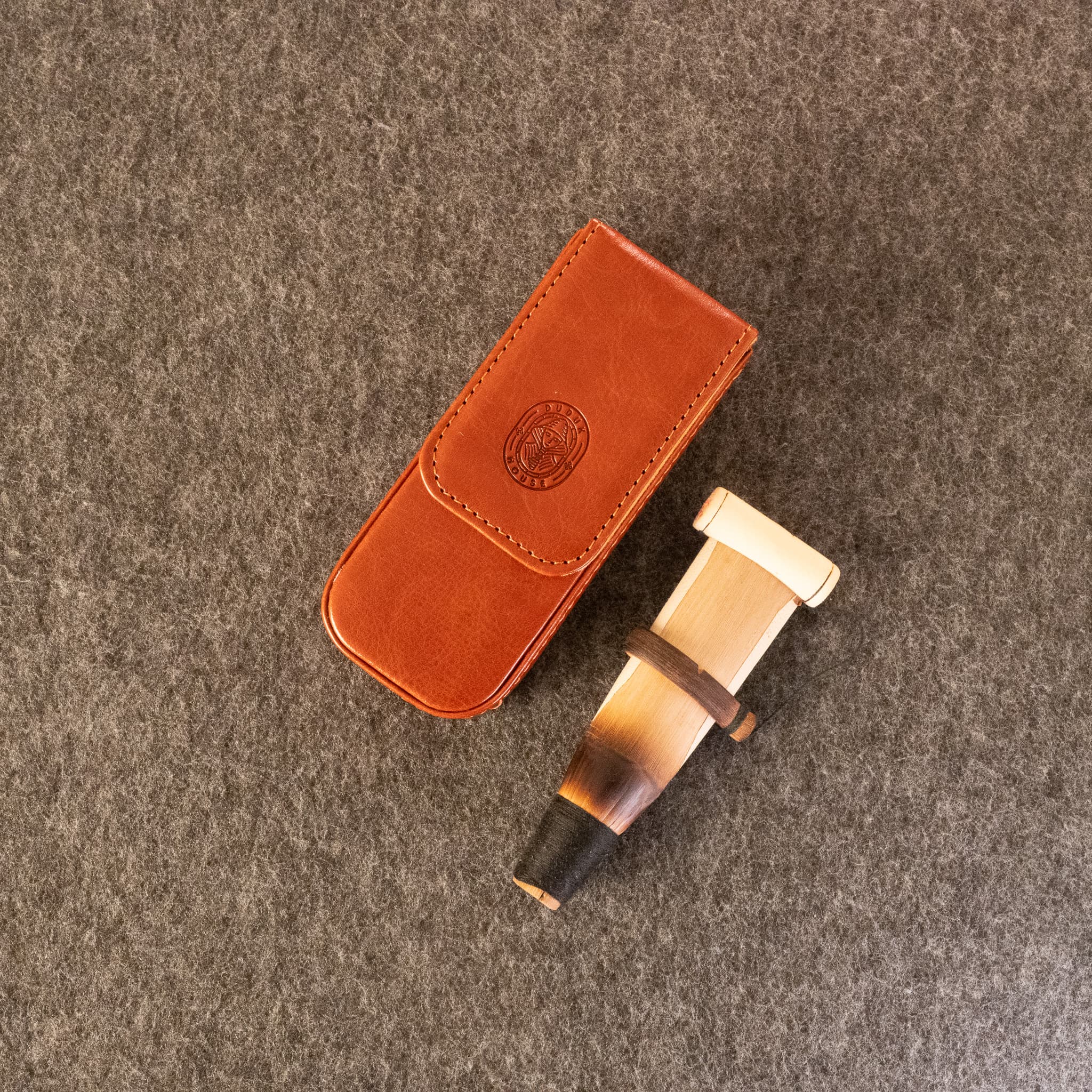 A handcrafted duduk reed, displayed next to its brown leather case on a textured gray felt background.