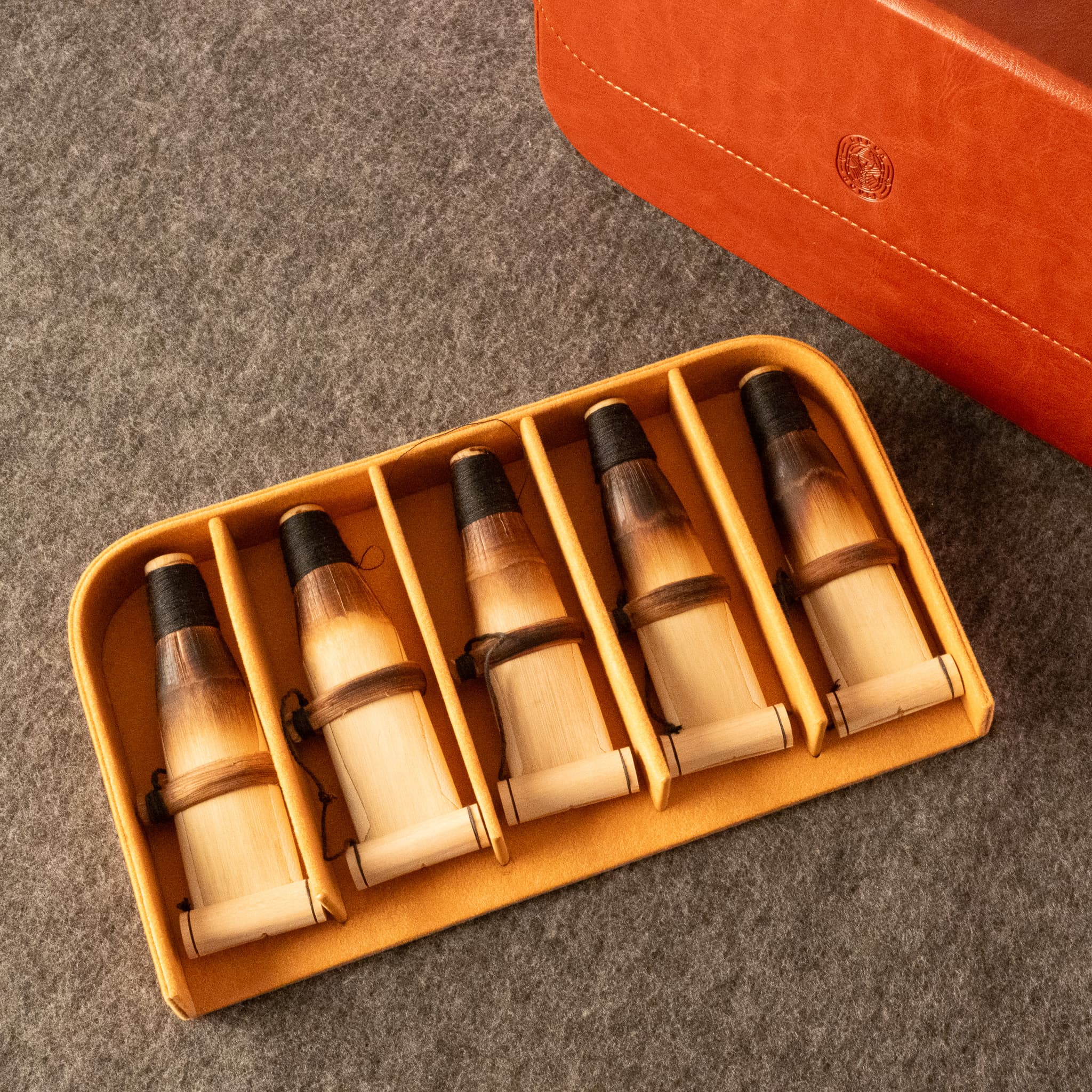 Five natural duduk reeds, displayed in a yellow-orange divided reed case on a gray felt surface, with part of an orange leather case with Dudukhouse logo visible in the upper right.